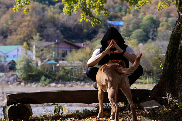 Man in prayer pose with dog by riverside. Young man in black hoodie sits cross-legged on wooden bench by river, hands in prayer, brown dog beside him, autumn foliage and mountains in background.