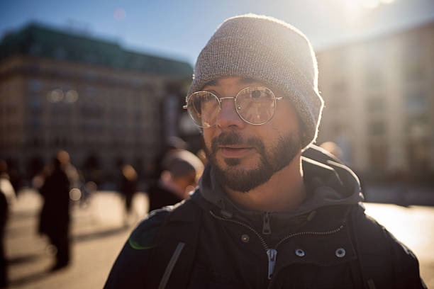 A man wearing a beanie and glasses stands in an urban environment, with sunlight illuminating his face. The background features blurred figures, suggesting a busy atmosphere. This image captures the essence of city life and can be used in contexts related to travel, lifestyle, or urban exploration.