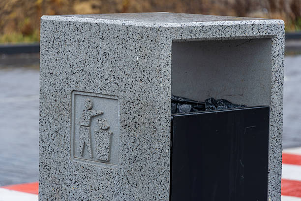 A sturdy trash bin made of stone is placed in a park, showing a symbol for proper waste disposal and filled with black trash bags