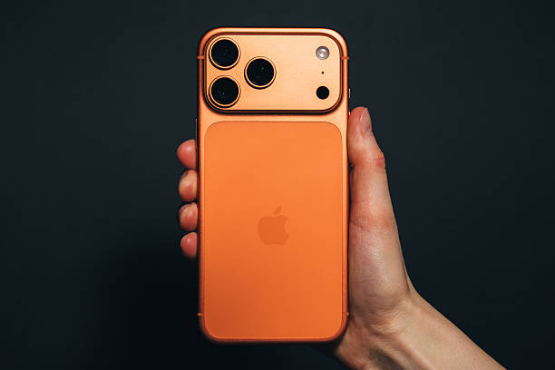 Krakow, Poland - November 6, 2025. Close-up editorial image shows a hand holding the latest Apple iPhone 17 Pro Max in Cosmic Orange, against a black background, highlighting the phone’s unique colour and camera design. The shot captures current consumer technology trends and the distinct design choices of Apple devices. This editorial photo is suitable for articles, reviews, and technology news related to smartphones, Apple products, or mobile innovation.