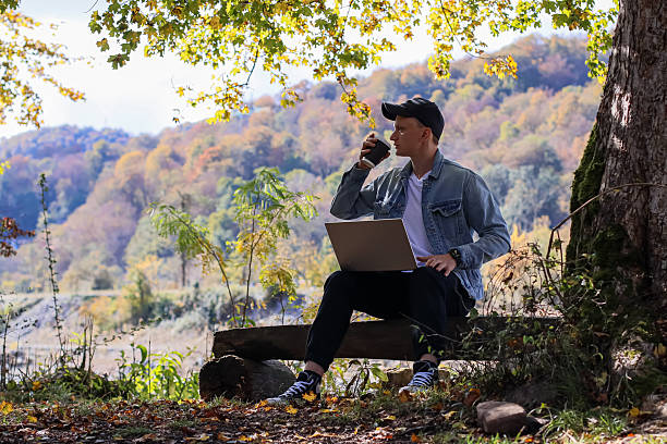 istock Young man drinks coffee at laptop on outdoor bench in fall woodland setting with valley backdrop. 2245999614