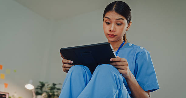 istock Young Asian female nurse in blue scrubs sitting on a bed while using a digital tablet. Nurse lifestyle concept. 2246005467