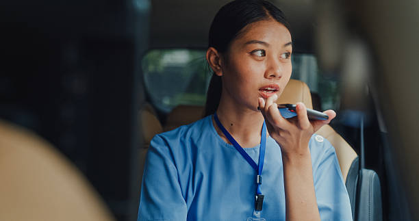 Young Asian female nurse in blue scrubs sitting in car, using a voice command feature on her phone. Nurse sustainable lifestyle concept.