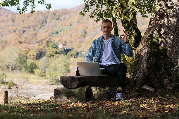 istock Man showing OK gesture seated with laptop under tree in colorful fall landscape 2246013667