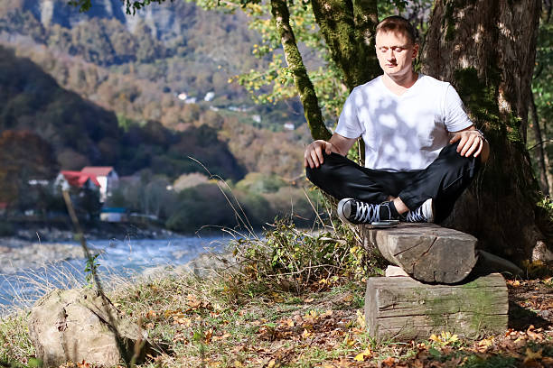 istock Man seated with hands on thighs by tree facing mountainous river 2246013938