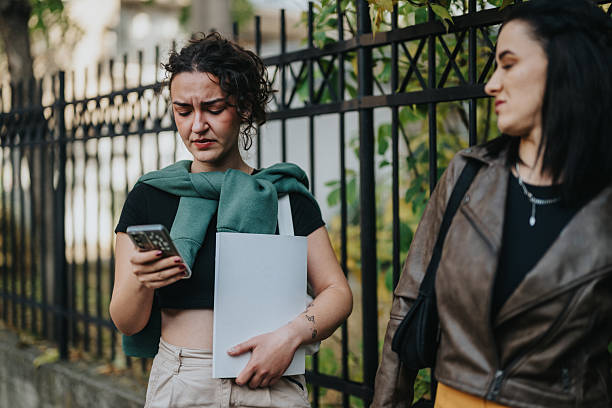 istock Two women walk along a fence-lined street, one focused on her phone while carrying a folder 2246021092