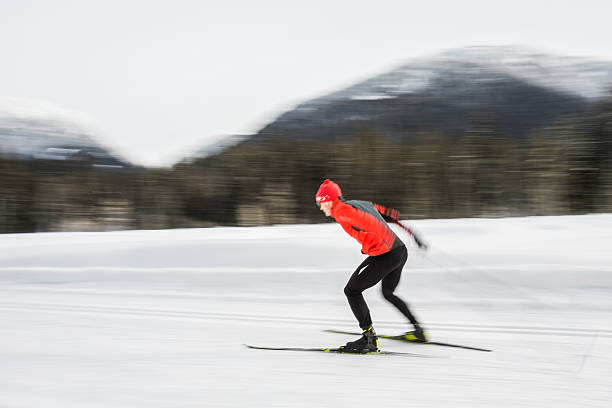 A skier in bright red gear descends swiftly down a snowy slope, surrounded by tall, snow-covered trees. The dynamic motion and wintry forest setting evoke a sense of adventure and exhilaration. The skier's vibrant outfit contrasts against the white snow, emphasizing the lively energy of this outdoor winter sport.