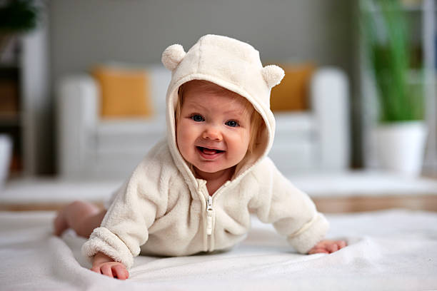 istock Cute Caucasian baby girl with blue eyes makes a funny grimace or playful expression while lying on her tummy in a cozy beige fleece jumpsuit with bear ears. 2246029820