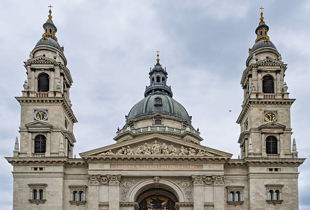 Budapest, HungaryThe image captures St. Stephen's Cathedral, Pest, Hungary, showcasing its prominent Gothic architecture. The cathedral's tall, ornate spires and façade with intricate stone carvings dominate the frame, viewed from street level in the urban setting of the city centre. The beige sandstone structure contrasts against a cloudy grey sky, with subtle greenery and modern elements like a low wall in the foreground. No people are present, emphasizing the building's grandeur. The mood is majestic and timeless, evoking historical significance. This image suits commercial uses such as travel guides highlighting European landmarks, architectural publications on Gothic design, tourism promotions for Vienna, historical documentaries, or editorial content on cultural heritage sites.