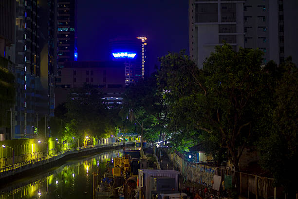Bunruang Watergate canal in Bangkok at night seen from Thanin Asol with view to east. Promenade at right side is under repairing constructions. In background is office building of Italian Thai construction company. Along canal are trees and buildings