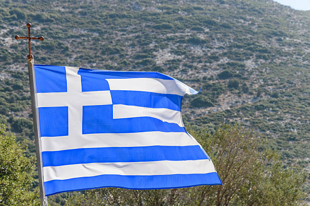 The national flag of Greece waves on a cross-topped pole against a backdrop of Mediterranean hillside vegetation. Featuring nine horizontal blue and white stripes and a white cross on a blue canton, the flag symbolizes Greek identity and Orthodox heritage. The natural setting highlights the country’s rugged terrain and cultural pride.