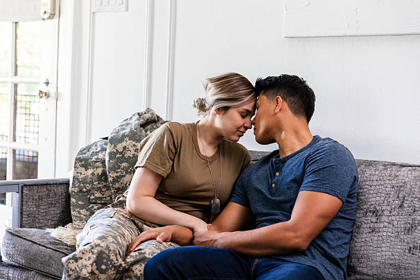 A couple shares an intimate moment on a sofa, one person in military uniform, holding hands and resting foreheads together.