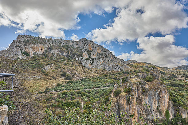 Rocky mountain terrain featuring rugged limestone cliffs and green scrubland under a blue sky
