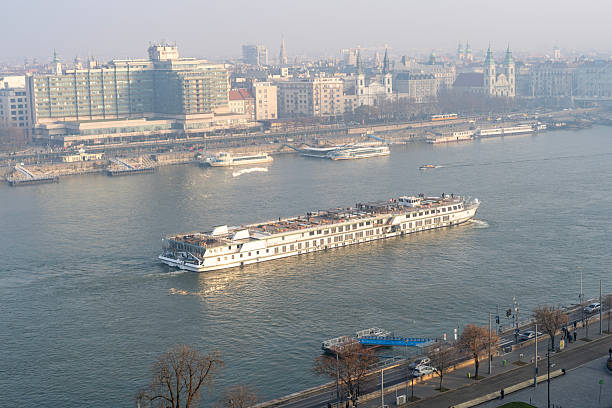 A large river cruise ship sails along a calm urban waterfront with historic and modern buildings in the background. Soft winter light and light haze create a peaceful atmosphere over the cityscape, while the vessel reflects gently on the water. The scene evokes travel, exploration, and tranquil riverfront scenery.