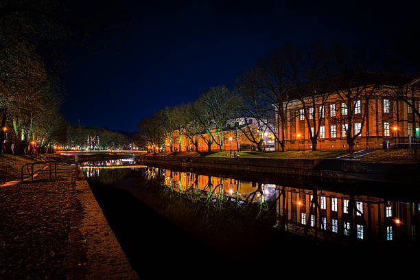 Beautiful night photograph of the Aurajoki River in Turku, Finland. The calm water reflects city lights and autumn colors, creating a peaceful and atmospheric urban scene.