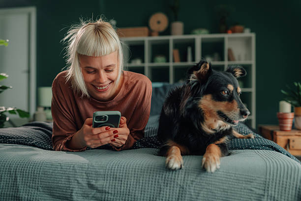 Woman smiling, petting dog while using phone on bed