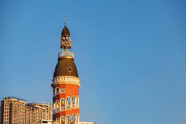 Red brick tower with a dark dome stands against blue sky. illuminated golden sunlight. Modern buildings are visible in the background. Showcasing a blend of architectural styles. Georgia, Batumi