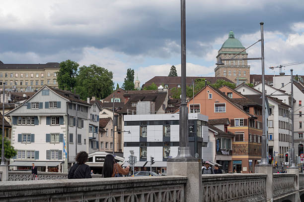 Bridge in Zurich spans the Limmat River with the ETH Zurich dome visible in the background. The scene includes diverse architectural styles, with pastel-colored, gabled-roof buildings lining the river. A cloudy summer sky looms above. Pedestrians, including men and women, walk along the bridge in casual attire. The area is lively, with cars and people moving through a busy urban setting. The ETH dome, a landmark in Switzerland, stands prominently, adding historical context to the modern environment.