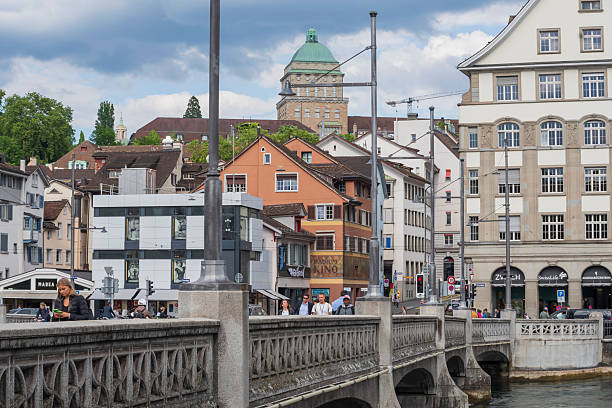 Bridge in Zurich spans the Limmat River with the ETH Zurich dome visible in the background. The scene includes diverse architectural styles, with pastel-colored, gabled-roof buildings lining the river. A cloudy summer sky looms above. Pedestrians, including men and women, walk along the bridge in casual attire. The area is lively, with cars and people moving through a busy urban setting. The ETH dome, a landmark in Switzerland, stands prominently, adding historical context to the modern environment.