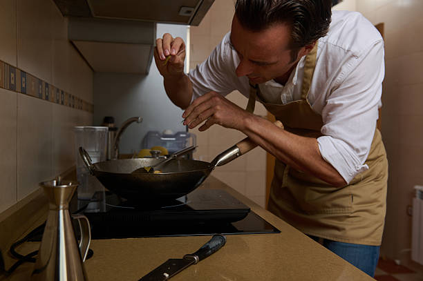 A man in an apron leaning over a frying pan on a stove, concentrating on cooking in a warm, home kitchen setting.
