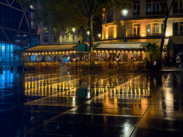This image is a dynamic night photograph taken in the Beaubourgarea of Paris, specifically in the square facing the Centre Pompidou, featuring the iconic Café Beaubourg. The scene is dominated by the dark, rain-slicked pavement which creates dramatic, bright yellow reflections of the café's warm string lights.The café is bustling with patrons visible inside and under a covered outdoor section, providing a sense of vibrant urban life despite the damp conditions. The foreground is filled with empty wicker chairs, adding a slightly tranquil note to the busy scene. Classic Parisian buildings are illuminated in the background, contrasting with the modern architecture and blue lighting of the nearby Centre Pompidou building visible on the far left. The image captures a quintessential, atmospheric Parisian evening in a lively cultural hub.