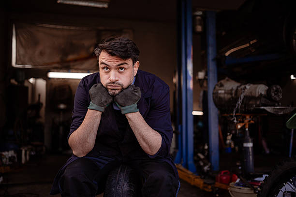 A mechanic sits on a tire in a workshop, looking thoughtful with hands on his cheeks amid tools and machinery.