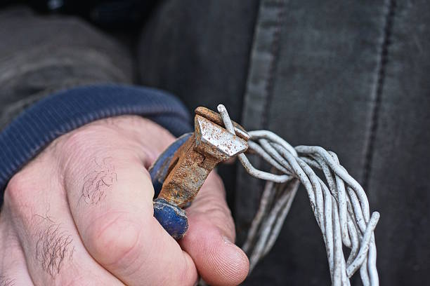 A man holds a rusty wire cutter in one hand, which he uses to cut off white aluminum wire on the street.