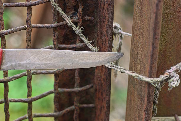 A single iron knife blade cuts a gray rope on a rusty brown post and a metal mesh fence on a summer street