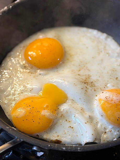 istock Full frame image of three merged, sunny side up fried eggs seasoned with ground black pepper, frying in non-stick frying pan, one runny broken orange yolk, steam rising, elevated view 2246084147