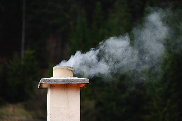 White smoke gently rises from a chimney surrounded by dense green forest, creating a calm and rustic atmosphere. The image evokes a sense of warmth, solitude, and connection with nature, contrasting the man-made structure with the natural background.