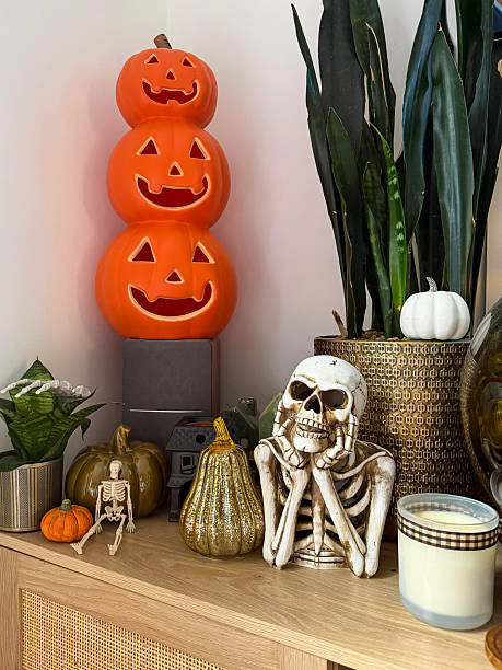 istock Close-up image Halloween decoration display arranged on wooden sideboard, stack of orange Jack O'lanterns on speaker, glazed china gourds and pumpkin, plastic skeleton, black model house, skeleton chin in hands, candle, potted plants, focus on foreground 2246085788