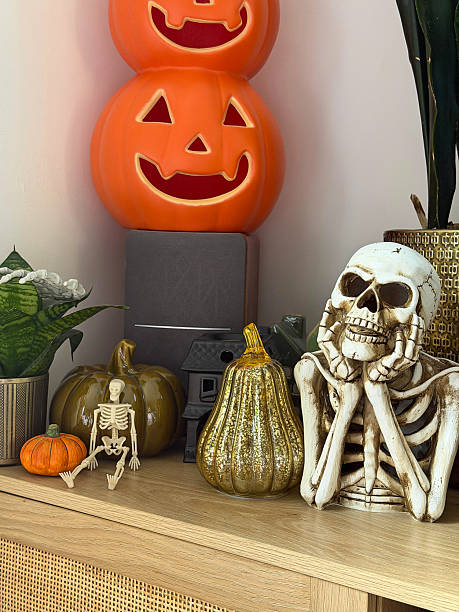 Close-up image Halloween decoration display arranged on wooden sideboard, stack of orange Jack O'lanterns on speaker, glazed china gourds and pumpkin, plastic skeleton, black model house, skeleton chin in hands, candle, potted plants, focus on foreground