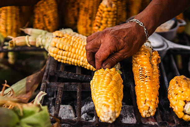 Green corn cobs roasting over charcoal, with rising smoke and golden texture. A typical Colombian street scene full of aroma, texture, and local tradition.