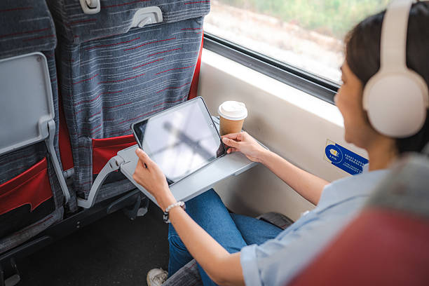 A beautiful Asian woman wearing headphones uses her digital tablet and enjoys a coffee on a train ride.