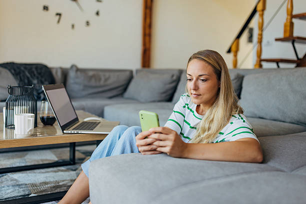 istock A smiling woman casually leans on a sofa, happily interacting with her mobile phone while an open laptop sits in front of her 2246125599