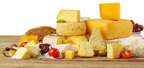Various types of cheese are arranged on a wooden table alongside fruits and bread.