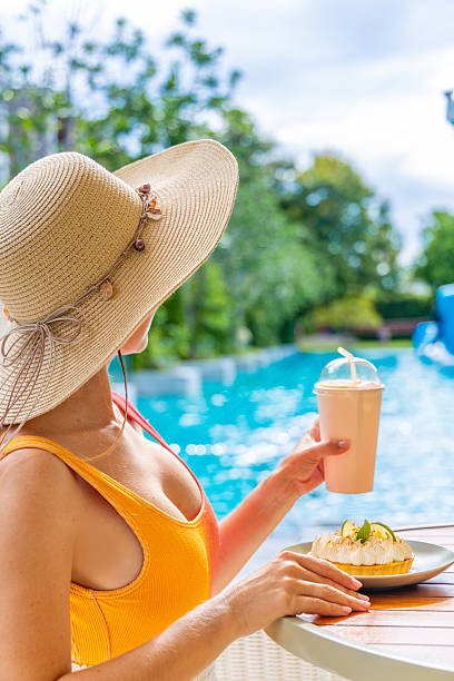Woman in sun hat and swimsuit enjoys summer poolside with smoothie and