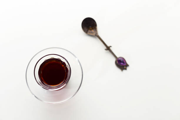 A warm beverage is placed in a clear glass next to a silver spoon on a white surface.