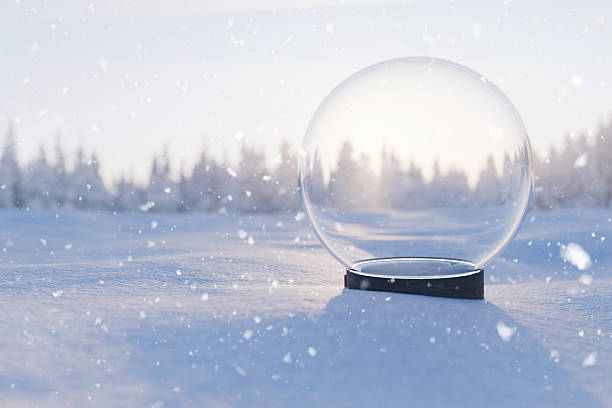 Empty snow globe on snowy ground with blurred winter forest at sunset light. Clear snow globe set in bright winter landscape with falling snow and sun glare. Place for product. Christmas creative idea