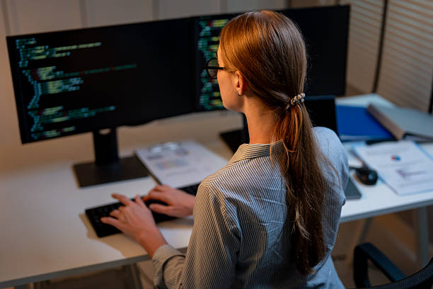 istock Rear view of young female software engineer working at night in office. Programmer IT technician looking at computer, creating solutions and develop technology security in workplace during evening. 2246132917