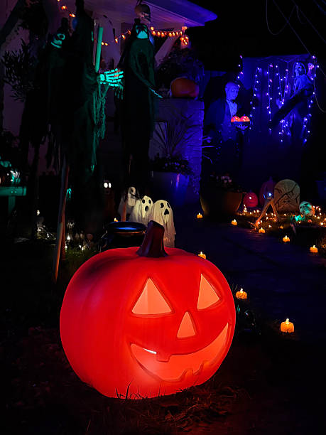 istock Image of front garden halloween scene with plastic jack-o’-lantern pumpkin faces, scary skeletons, glowing pumpkins, ghosts, purple and green fairy lights 2246137103