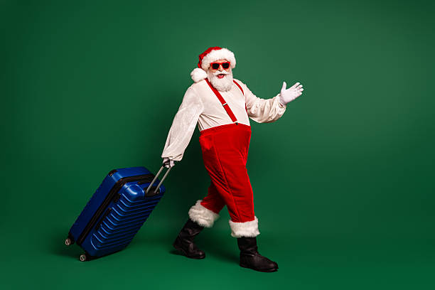 A cheerful Santa in a traditional red costume pulls a blue suitcase on a green studio backdrop, creating a playful holiday scene perfect for Christmas and travel themed campaigns.