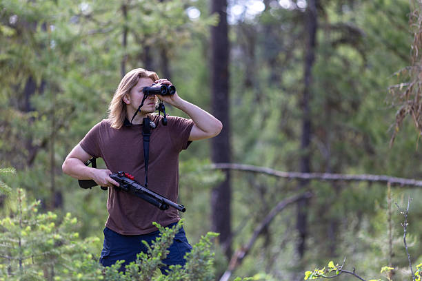 istock Young man observing terrain with binoculars while holding a shotgun ready. 2246139369