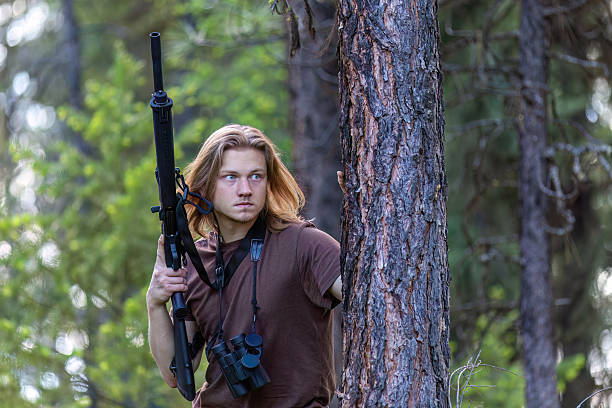 Young man, hunter, stands partially hidden behind tree trunk in deep forest. He holds a shotgun vertically in his hand and wears binoculars around his neck. Concept of hunting, scouting, or wilderness survival.