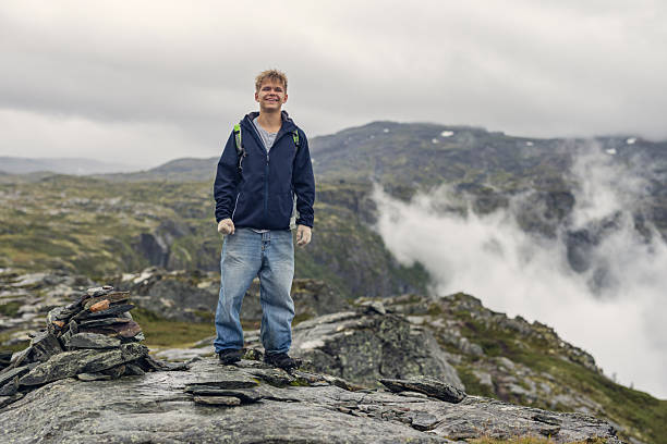 Teenage hiker stands alone on a rocky mountain summit overlooking a sea of clouds filling the fjord below. The hiker smiles at camera with the dramatic Norwegian mountain landscape where thick fog flows between peaks, creating an ethereal cloudscape. Mountain ridges emerge from the mist in layers.Shot with Canon R5 on an overcast summer day.
