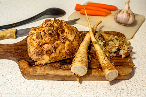 Halved celery root and parsley on olive wood cutting board, carrot and onion, spoon and knife on kitchen table. Preparation of meal.