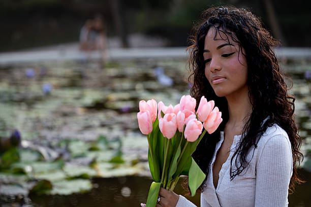 Thoughtful young woman holding a bouquet of pink tulips, looking down by a peaceful pond with water lilies