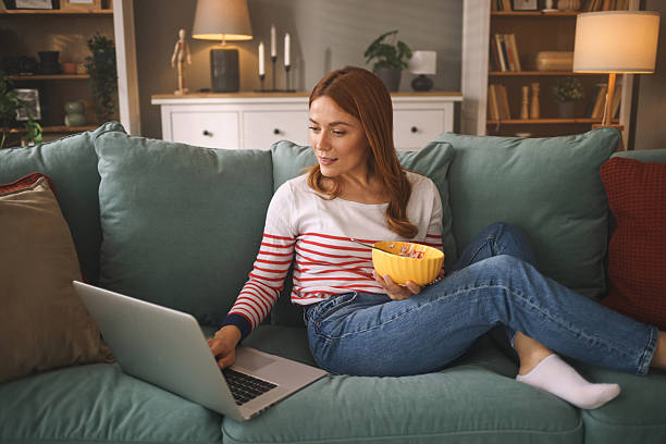 istock Young woman relaxing on sofa, eating and browsing internet on laptop 2246149846
