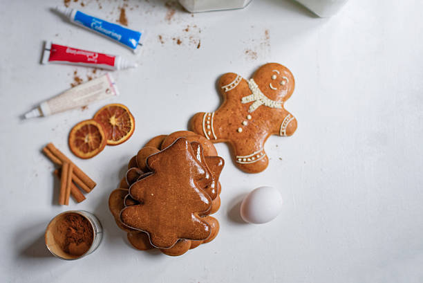 Minimal flat-lay of stacked gingerbread cookies and a smiling gingerbread man surrounded by icing, spices and an egg on a white background. Festive Christmas baking, clean composition, and copy space ideal for promotions or recipes.