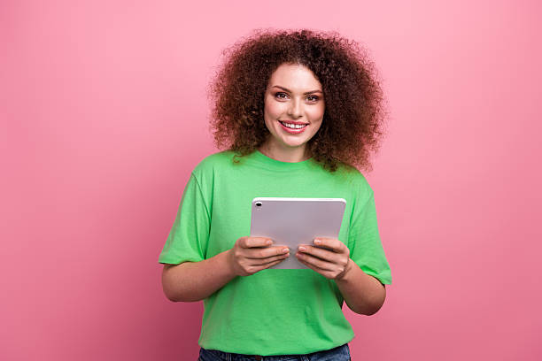 istock Young woman with curly brunette hair wearing a bright green tshirt smiling while using a tablet against a pink background 2246152563